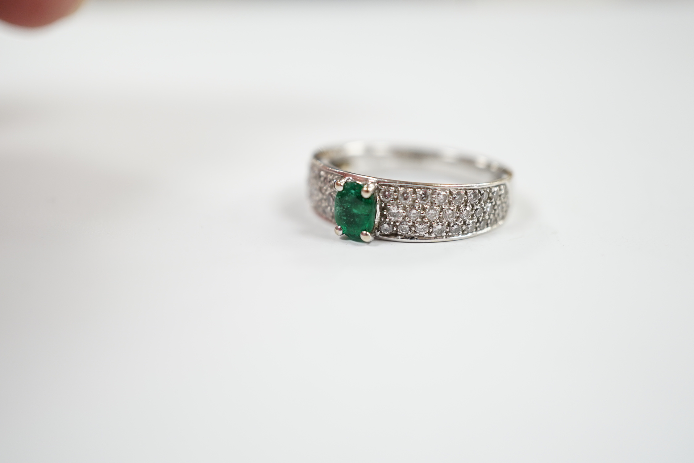 A modern 18ct white gold and single stone oval cut emerald set ring, with pave set diamond shoulders, size L/M, gross weight 4.3 grams.